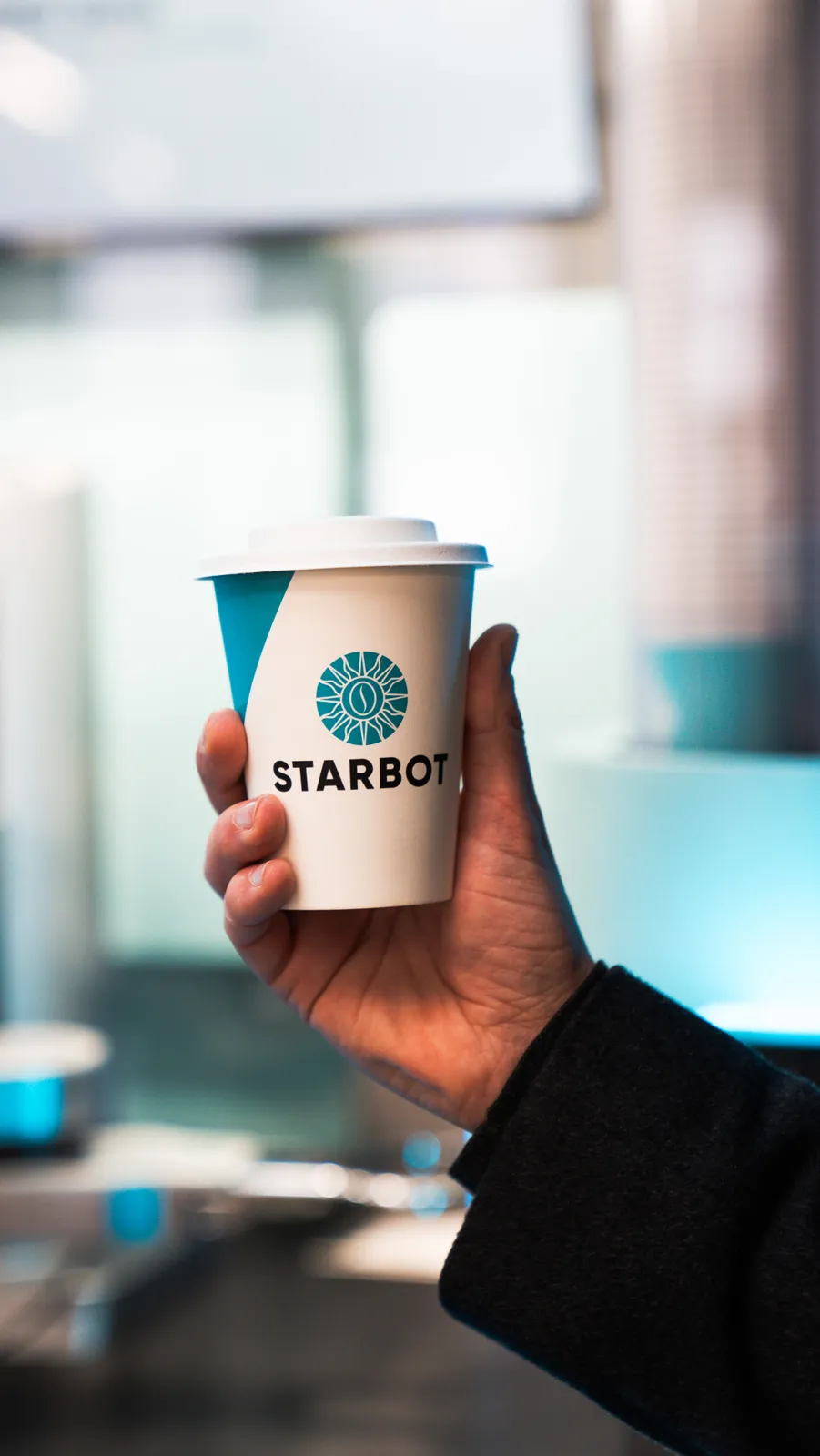 A branded STARBOT cup held in front of the coffee kiosk